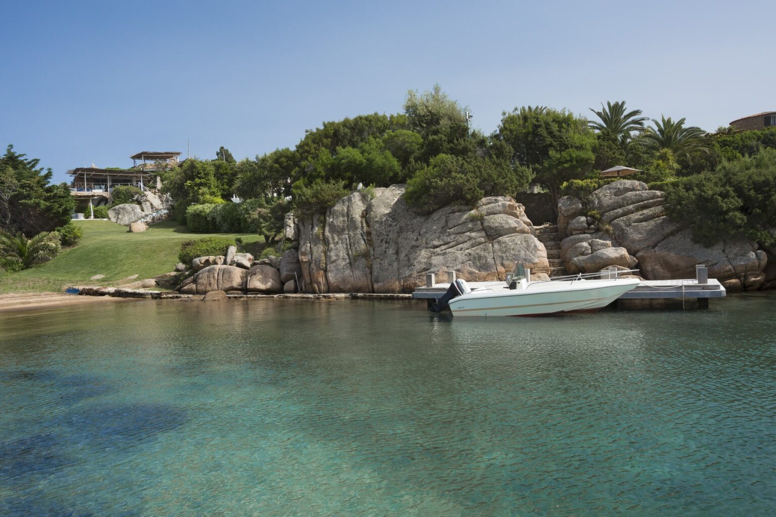 Scenic coastal view featuring clear turquoise waters, a white boat docked at a wooden pier, and lush greenery on rocky cliffs under a clear blue sky. Ideal for beach and nature enthusiasts. | Only on SopranoVillas