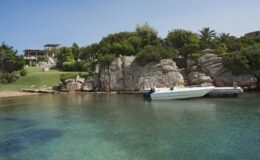 Scenic coastal view featuring clear turquoise waters, a white boat docked at a wooden pier, and lush greenery on rocky cliffs under a clear blue sky. Ideal for beach and nature enthusiasts.