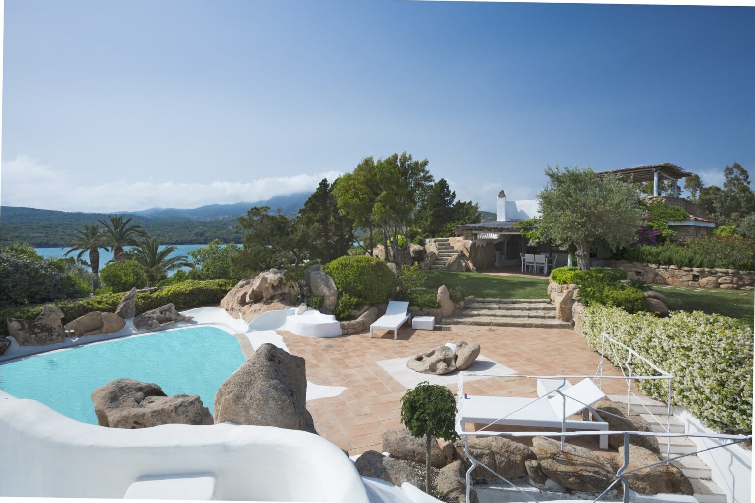 Luxurious villa with a swimming pool surrounded by natural stone, lush greenery, and palm trees, overlooking a serene lake and distant mountains in a sunny landscape. Perfect for relaxation and outdoor gatherings. | Only on SopranoVillas