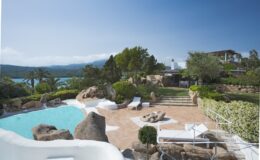 Luxurious villa with a swimming pool surrounded by natural stone, lush greenery, and palm trees, overlooking a serene lake and distant mountains in a sunny landscape. Perfect for relaxation and outdoor gatherings.