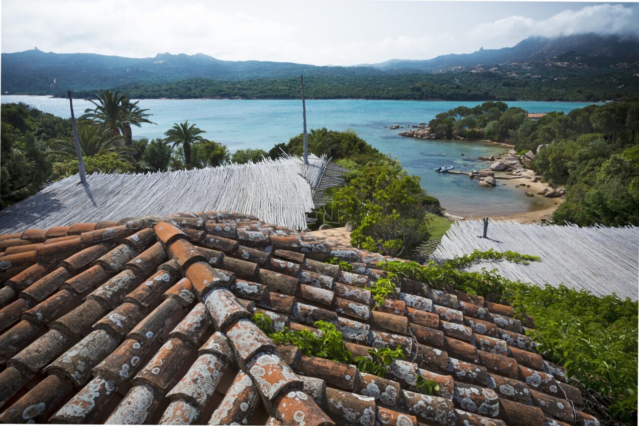 Scenic view of a coastal landscape featuring terracotta rooftops, lush greenery, and a turquoise sea with distant mountains under a partly cloudy sky.