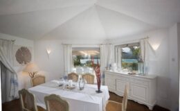 Bright and airy dining room with a white table set for a meal, elegant chairs, and large windows showcasing a scenic view. The room features soft lighting, decorative curtains, and a stylish sideboard, creating a welcoming atmosphere for dining.