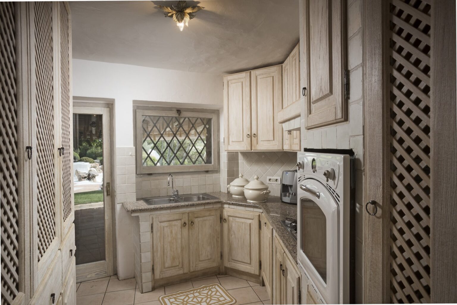 Cozy kitchen with light wood cabinets, granite countertops, and a window featuring decorative lattice. The layout is compact, showcasing modern appliances and a warm, inviting atmosphere. | Only on SopranoVillas