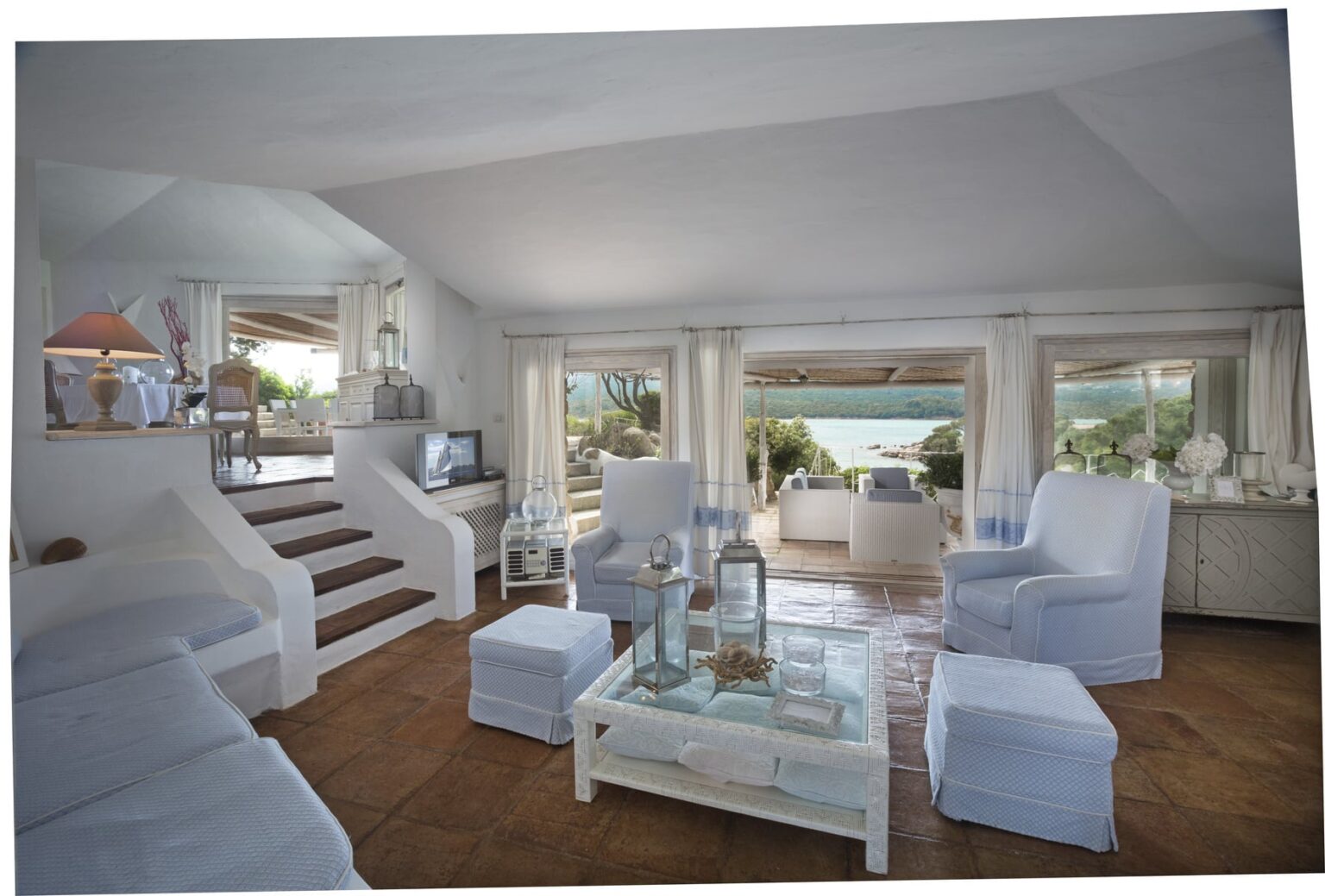 Bright and airy living room with white furniture, large windows showcasing a scenic view, and a cozy atmosphere. The room features a staircase, a glass coffee table, and decorative accents, creating a welcoming space for relaxation. | Only on SopranoVillas