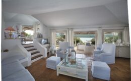 Bright and airy living room with white furniture, large windows showcasing a scenic view, and a cozy atmosphere. The room features a staircase, a glass coffee table, and decorative accents, creating a welcoming space for relaxation.