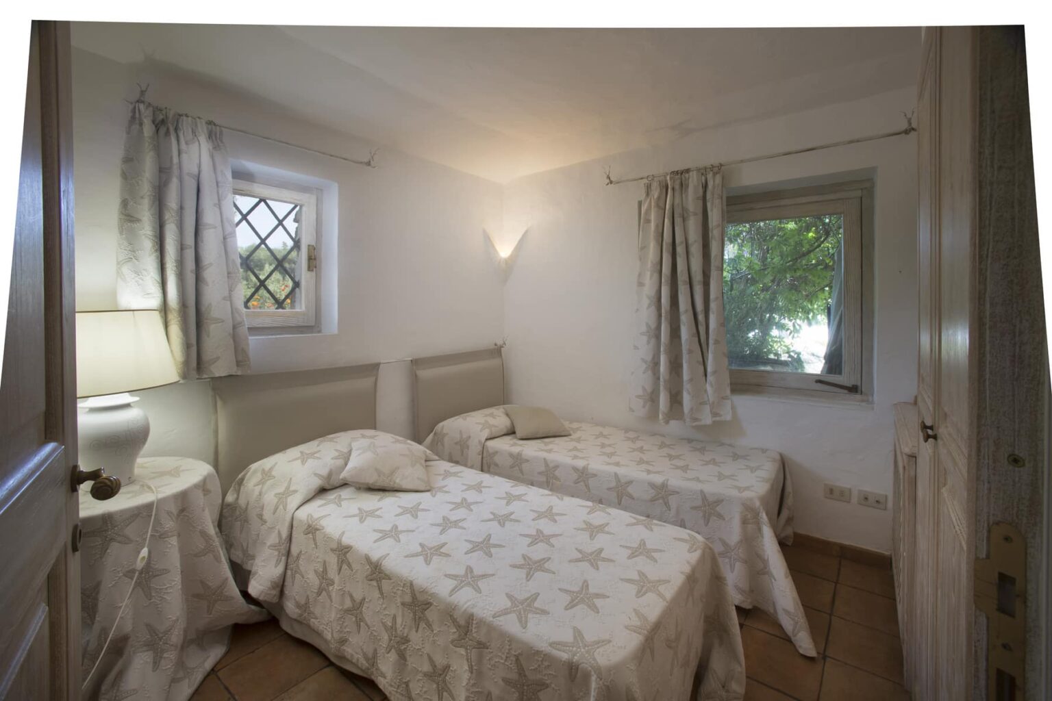 Cozy bedroom featuring two twin beds with star-patterned bedding, a bedside lamp, and natural light coming through a window with leafy views. The room has light-colored walls and soft curtains, creating a serene atmosphere. | Only on SopranoVillas