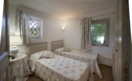 Cozy bedroom featuring two twin beds with star-patterned bedding, a bedside lamp, and natural light coming through a window with leafy views. The room has light-colored walls and soft curtains, creating a serene atmosphere.
