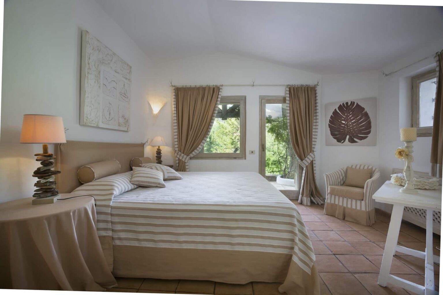 Cozy bedroom featuring a comfortable double bed with striped linens, warm beige curtains, a bedside lamp, and a small seating area. Natural light streams in through large windows, enhancing the serene ambiance. | Only on SopranoVillas