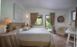 Cozy bedroom featuring a comfortable double bed with striped linens, warm beige curtains, a bedside lamp, and a small seating area. Natural light streams in through large windows, enhancing the serene ambiance.
