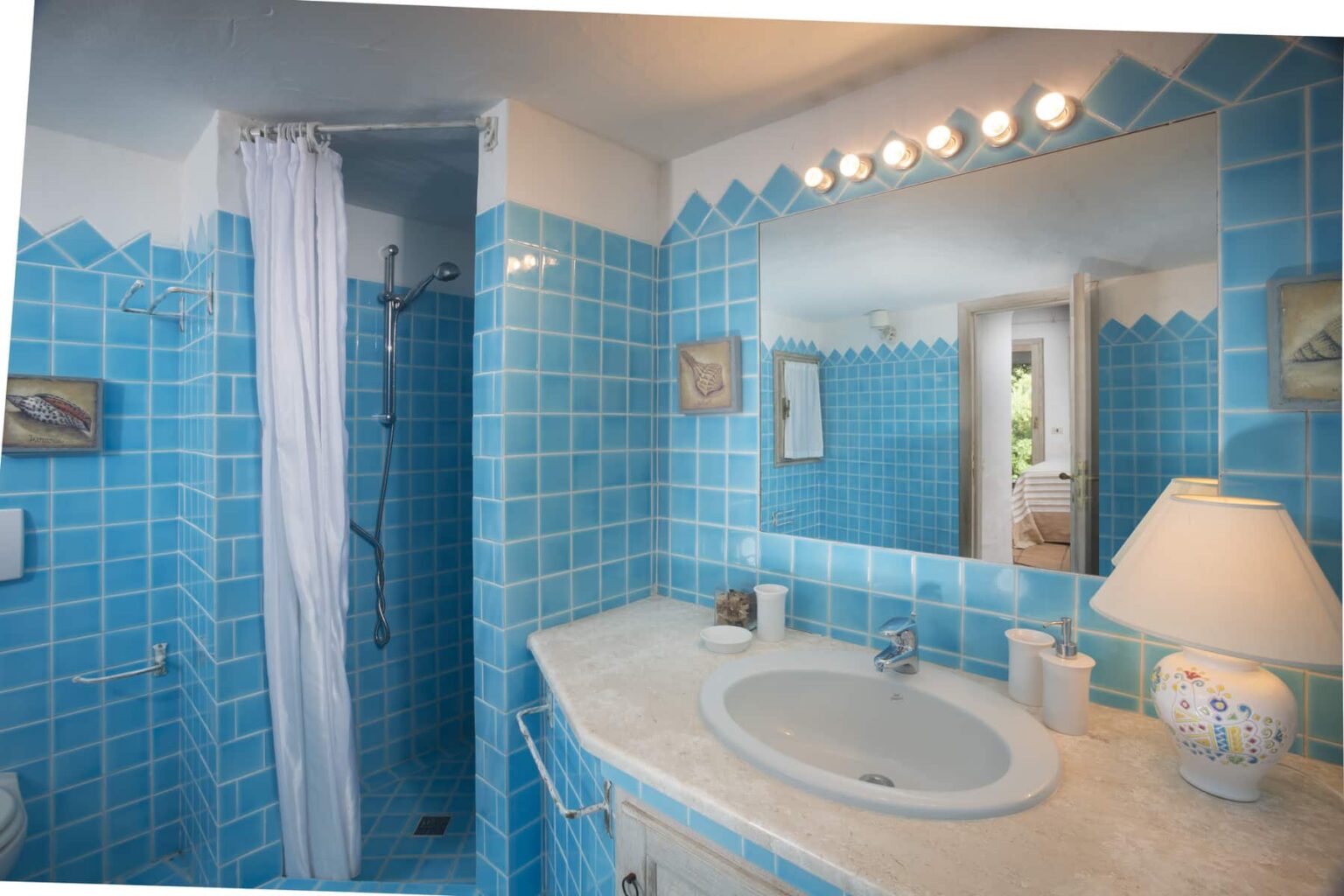 Bright and airy bathroom featuring turquoise tiled walls, a modern sink with a marble countertop, a shower area with a curtain, and a stylish mirror illuminated by overhead lights. A lamp and decorative elements enhance the inviting atmosphere. | Only on SopranoVillas