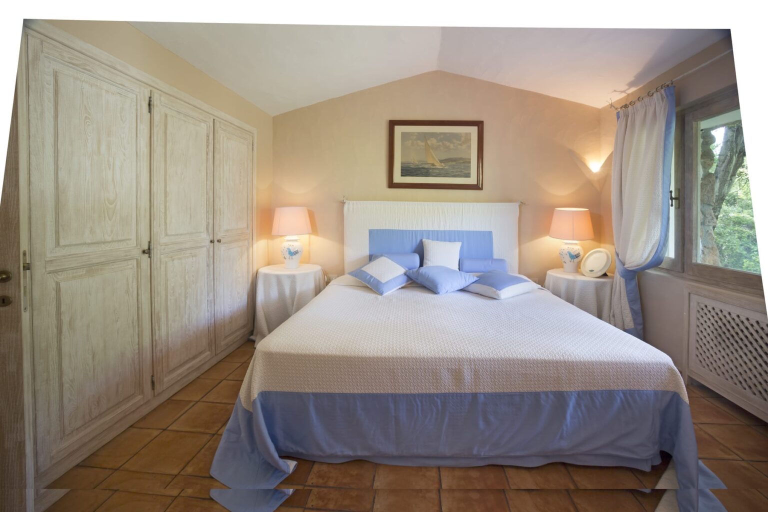Cozy bedroom featuring a king-sized bed with blue and white bedding, two bedside lamps, and large windows allowing natural light. The room includes a spacious wardrobe and a serene color palette, creating a relaxing atmosphere. | Only on SopranoVillas