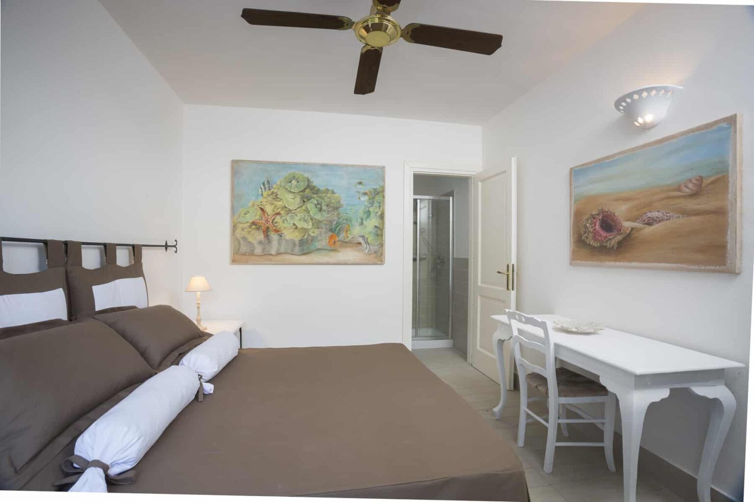 Cozy hotel room featuring a queen-sized bed with brown bedding, a white desk and chair, and colorful artwork on the walls. The room includes a ceiling fan and an en-suite bathroom. Ideal for travelers seeking comfort and style. | Only on SopranoVillas