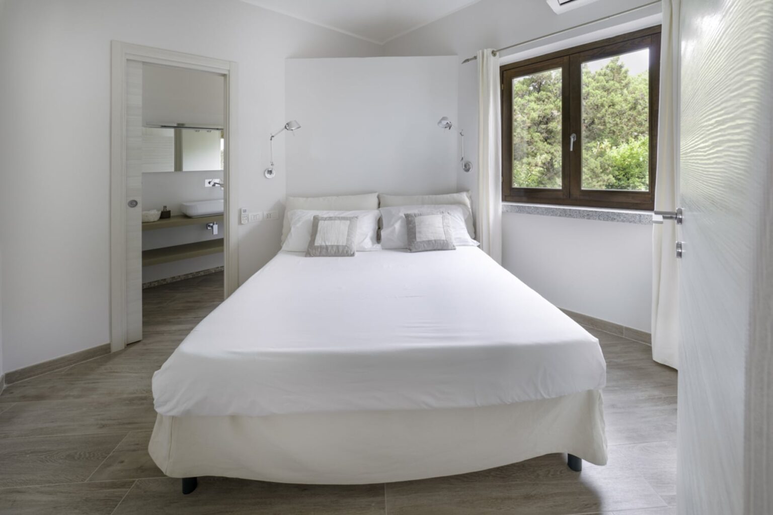 Bright and airy bedroom featuring a large bed with white linens and decorative pillows, complemented by modern wall sconces. A window offers a view of greenery outside, while a doorway leads to a stylish bathroom with minimalist decor. Ideal for relaxation and comfort. | Only on SopranoVillas