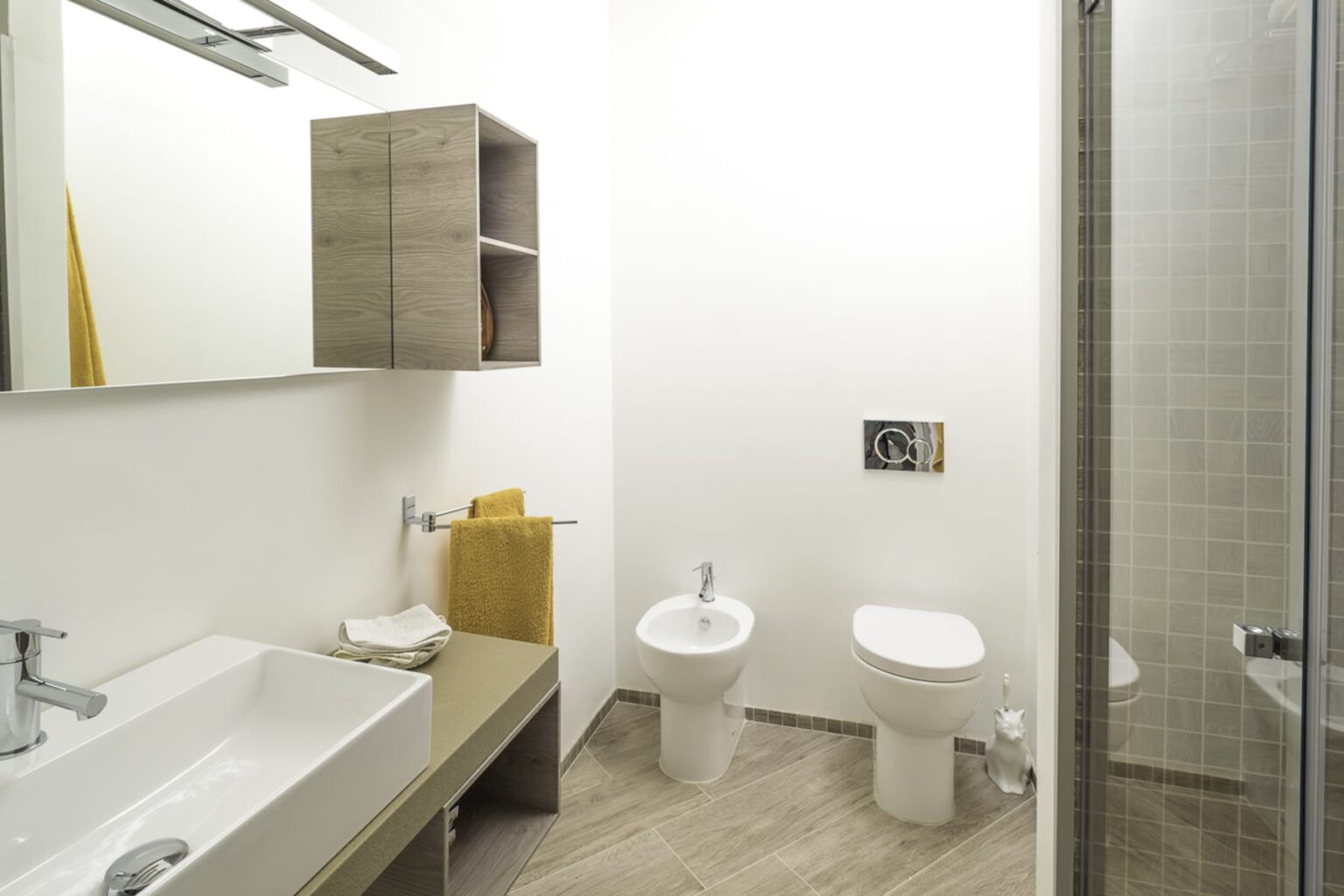 Modern bathroom featuring a spacious layout with a sink, bidet, toilet, and shower. The design includes wooden cabinetry and neutral-colored walls, creating a clean and minimalist aesthetic. | Only on SopranoVillas
