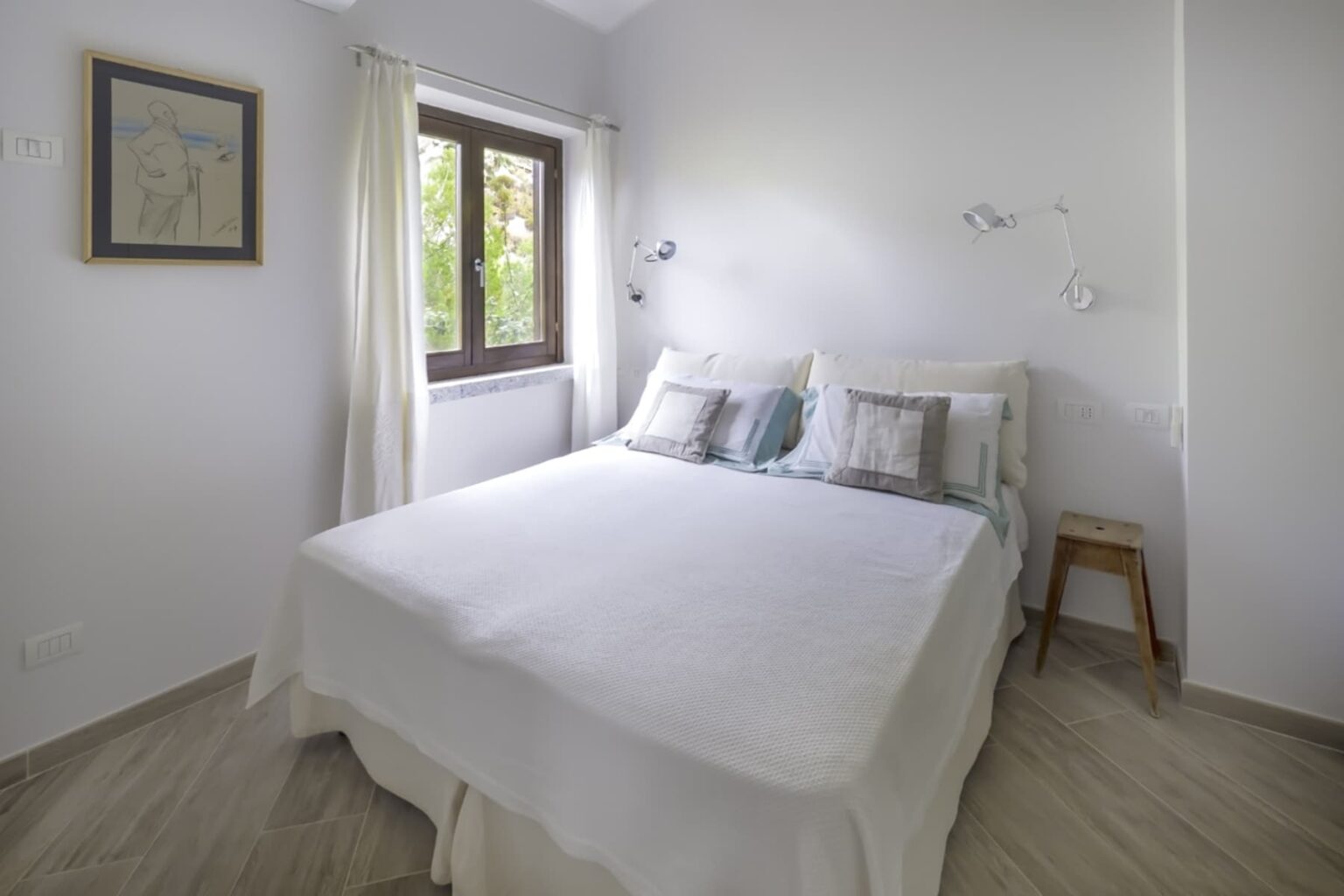 Cozy modern bedroom featuring a queen-size bed adorned with decorative pillows, light-colored bedding, and bedside wall lamps. A large window with sheer curtains allows natural light to illuminate the space, complemented by a framed artwork on the wall. The room has neutral-colored walls and stylish tile flooring, creating a serene and inviting atmosphere. | Only on SopranoVillas