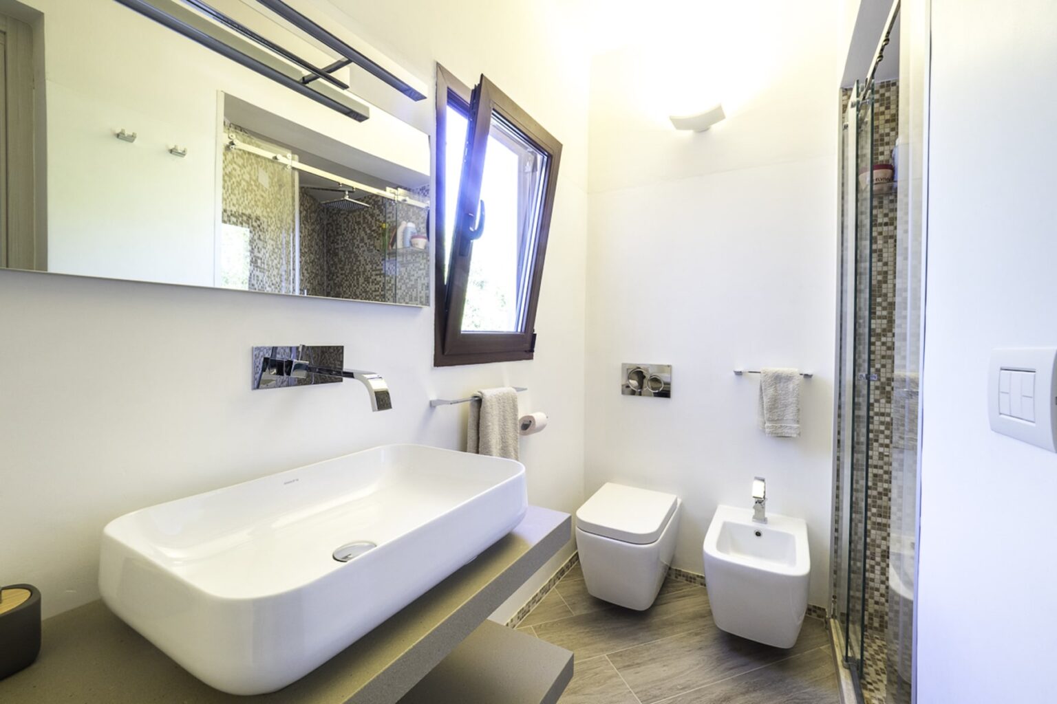 Modern bathroom featuring a stylish freestanding bathtub, bidet, and a large mirror. Natural light streams in through a window, highlighting the sleek design and neutral color palette. | Only on SopranoVillas