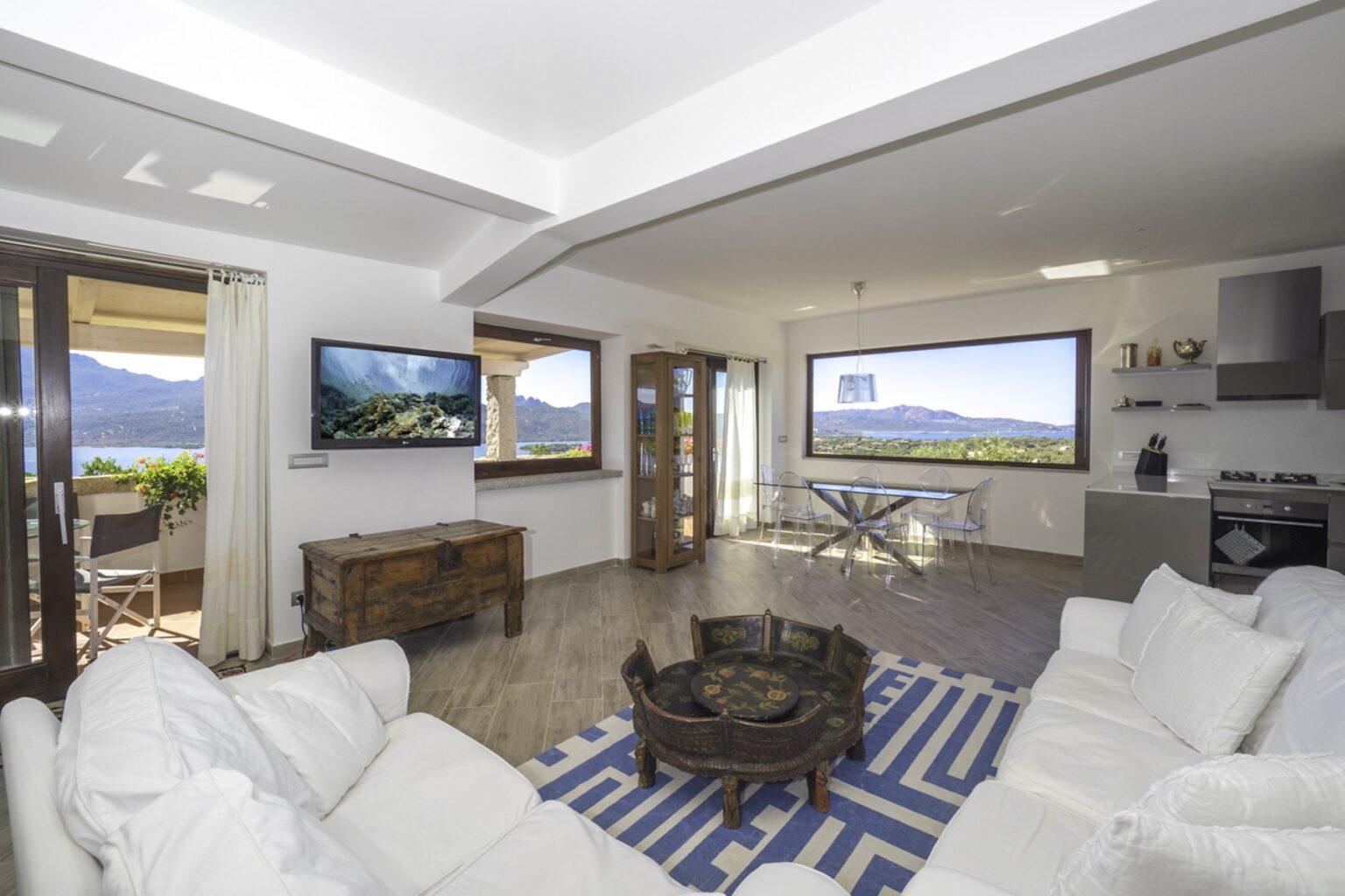 Modern living room with white sofas, a wooden coffee table, and large windows showcasing mountain views. Features a flat-screen TV and an open layout connecting to a dining area and kitchen. Bright, inviting atmosphere with natural light. | Only on SopranoVillas