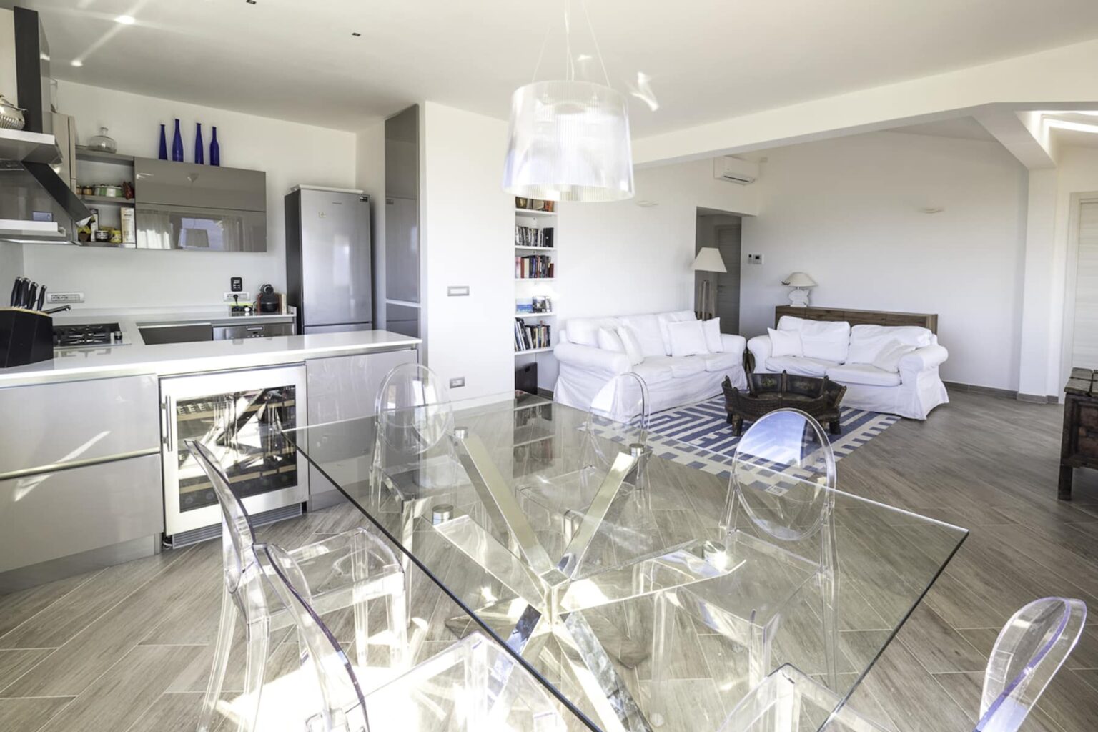 Modern open-concept living room and kitchen featuring a glass dining table, sleek appliances, and cozy seating arrangements, with natural light illuminating the space. | Only on SopranoVillas