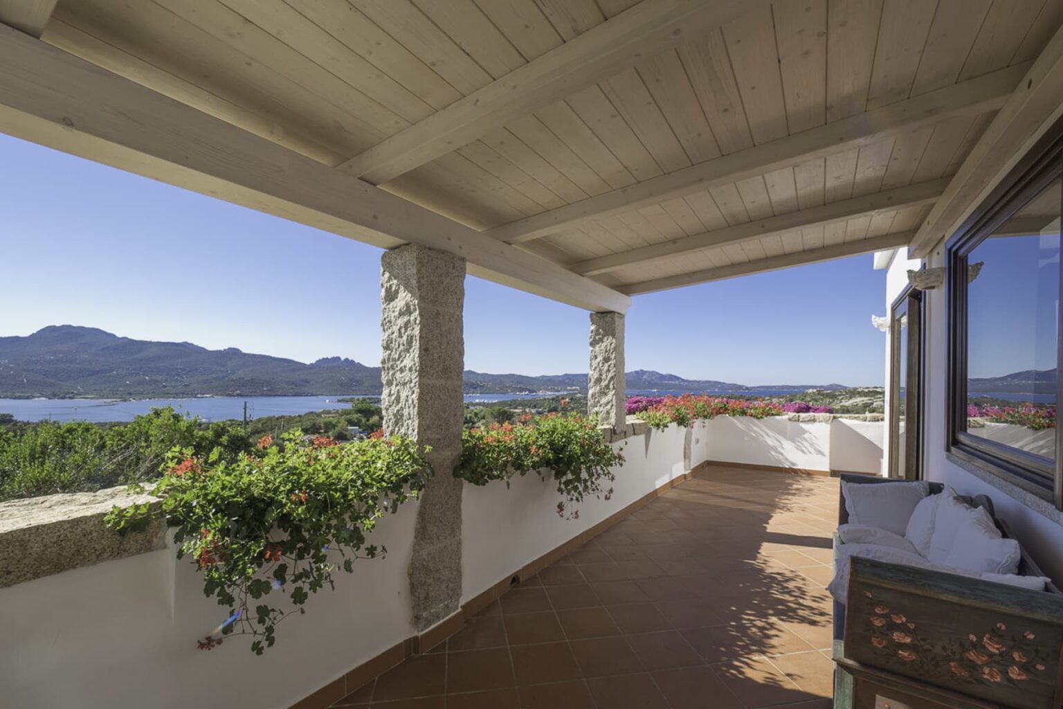 Spacious balcony with a wooden ceiling, featuring potted plants, offering a panoramic view of mountains and a serene lake under a clear blue sky. | Only on SopranoVillas