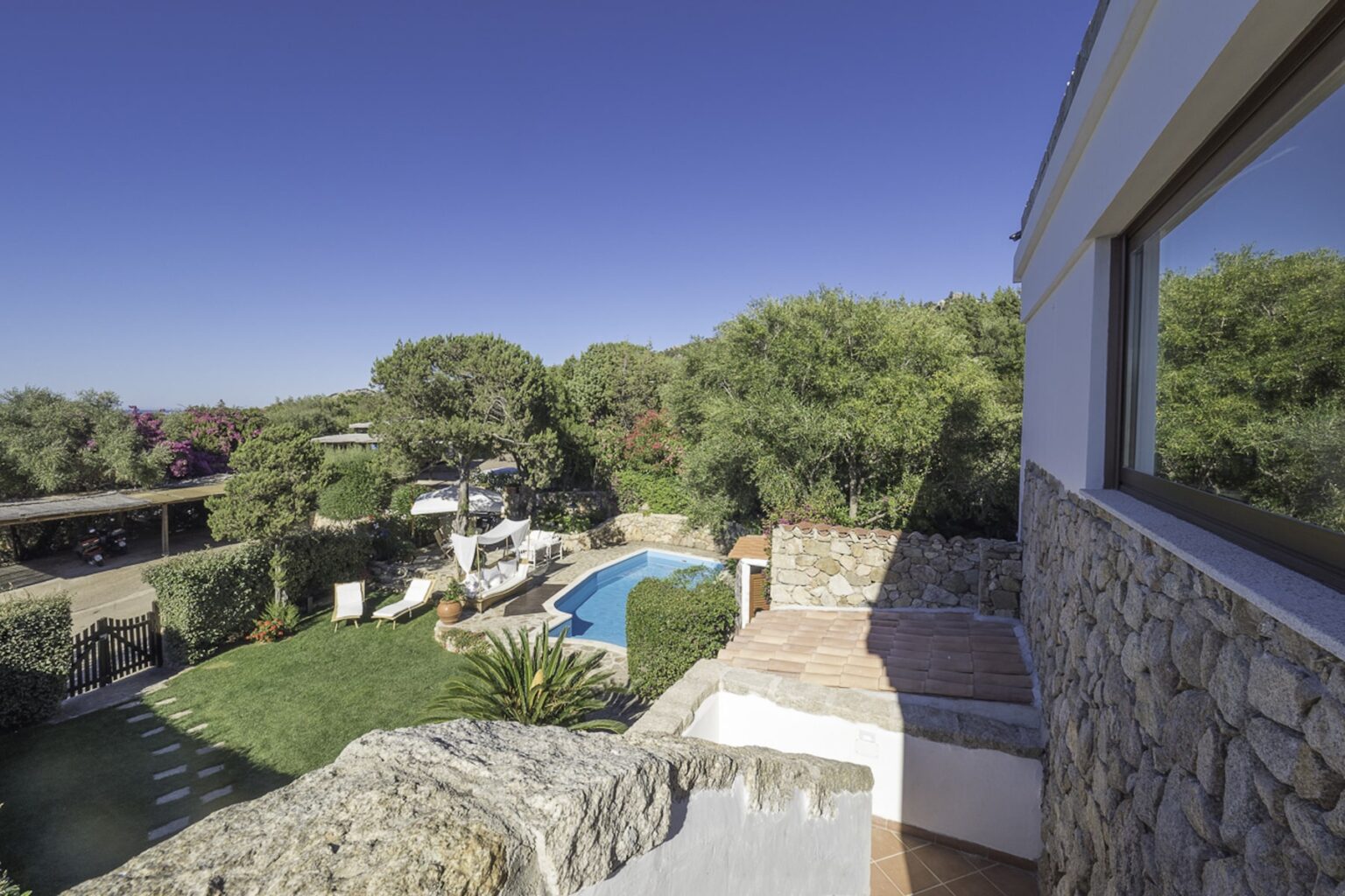 Scenic view of a landscaped garden with a blue swimming pool, surrounded by trees and stone pathways, captured from a balcony. | Only on SopranoVillas