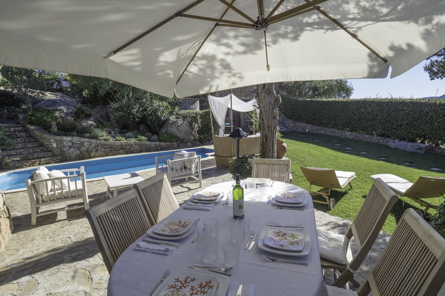 Outdoor dining area set for a meal, featuring a large table with plates and a bottle of wine, shaded by an umbrella, overlooking a swimming pool and lush garden. | Only on SopranoVillas