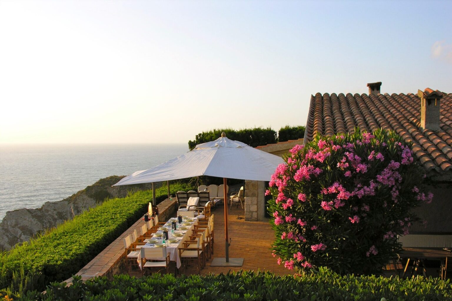 Scenic outdoor dining area overlooking the ocean, featuring a large table set for a meal under a white umbrella, surrounded by lush greenery and vibrant pink flowers, with a rustic house in the background. | Only on SopranoVillas