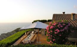 Scenic outdoor dining area overlooking the ocean, featuring a large table set for a meal under a white umbrella, surrounded by lush greenery and vibrant pink flowers, with a rustic house in the background.