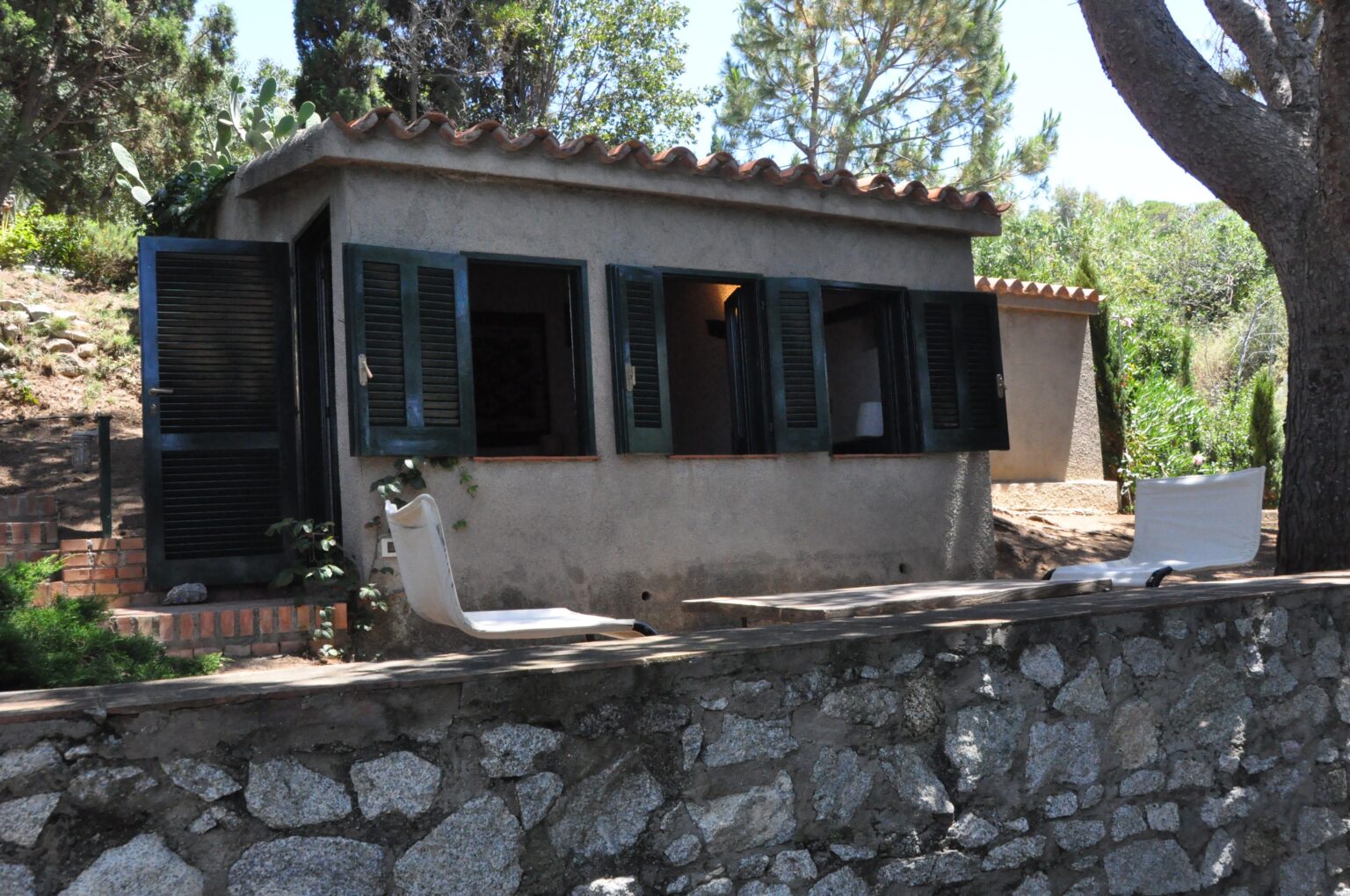 Charming rustic cabin with green shutters, surrounded by trees and plants, featuring a stone wall and wooden deck for outdoor relaxation. Ideal for nature retreats and cozy getaways. | Only on SopranoVillas