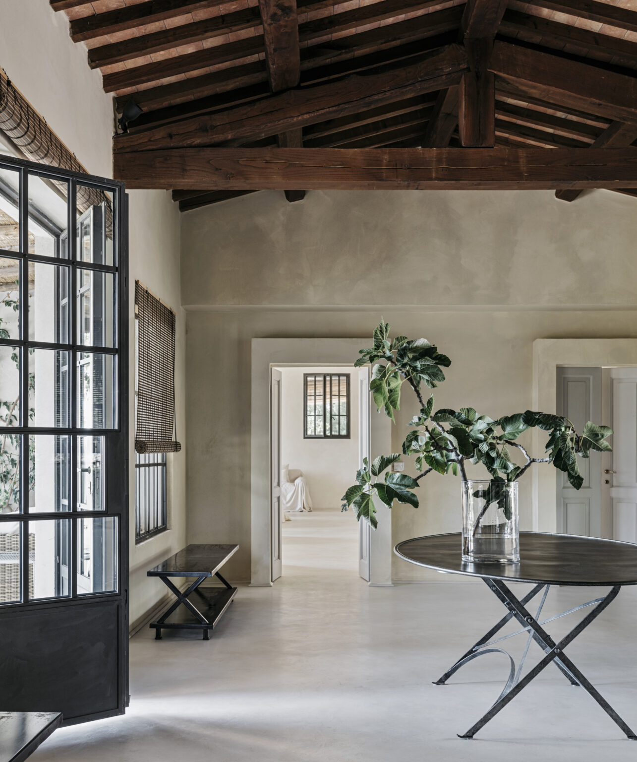 Modern interior design featuring a spacious room with high wooden beams, large windows, and a minimalist aesthetic. A round table with a glass vase and foliage is in focus, while an open doorway leads to another softly lit area. The neutral color palette and natural light create a serene atmosphere. | Only on SopranoVillas