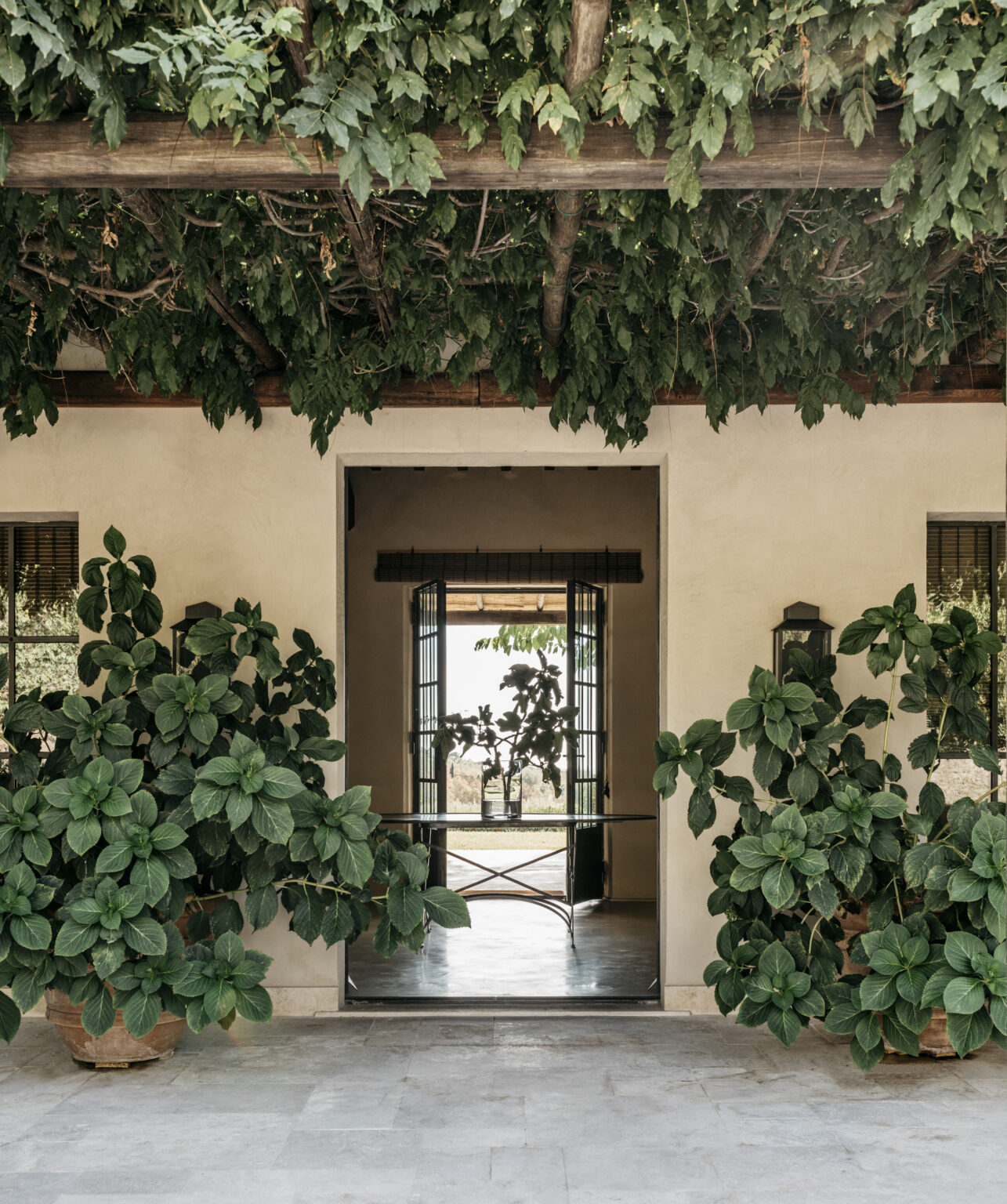 Charming entrance of a Mediterranean-style home featuring lush greenery, potted plants, and an inviting view through open doors, showcasing a serene outdoor landscape. | Only on SopranoVillas