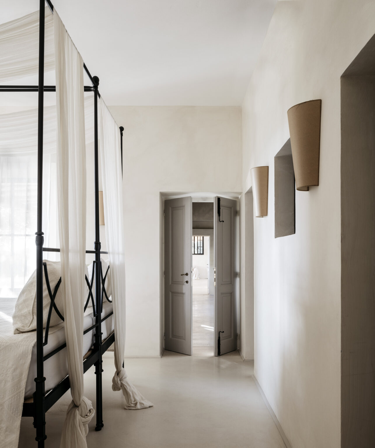 Modern bedroom interior featuring a black four-poster bed with sheer curtains, leading to a softly lit hallway with open doors and minimalist decor. The walls are painted in a light tone, enhancing the airy atmosphere of the space. | Only on SopranoVillas