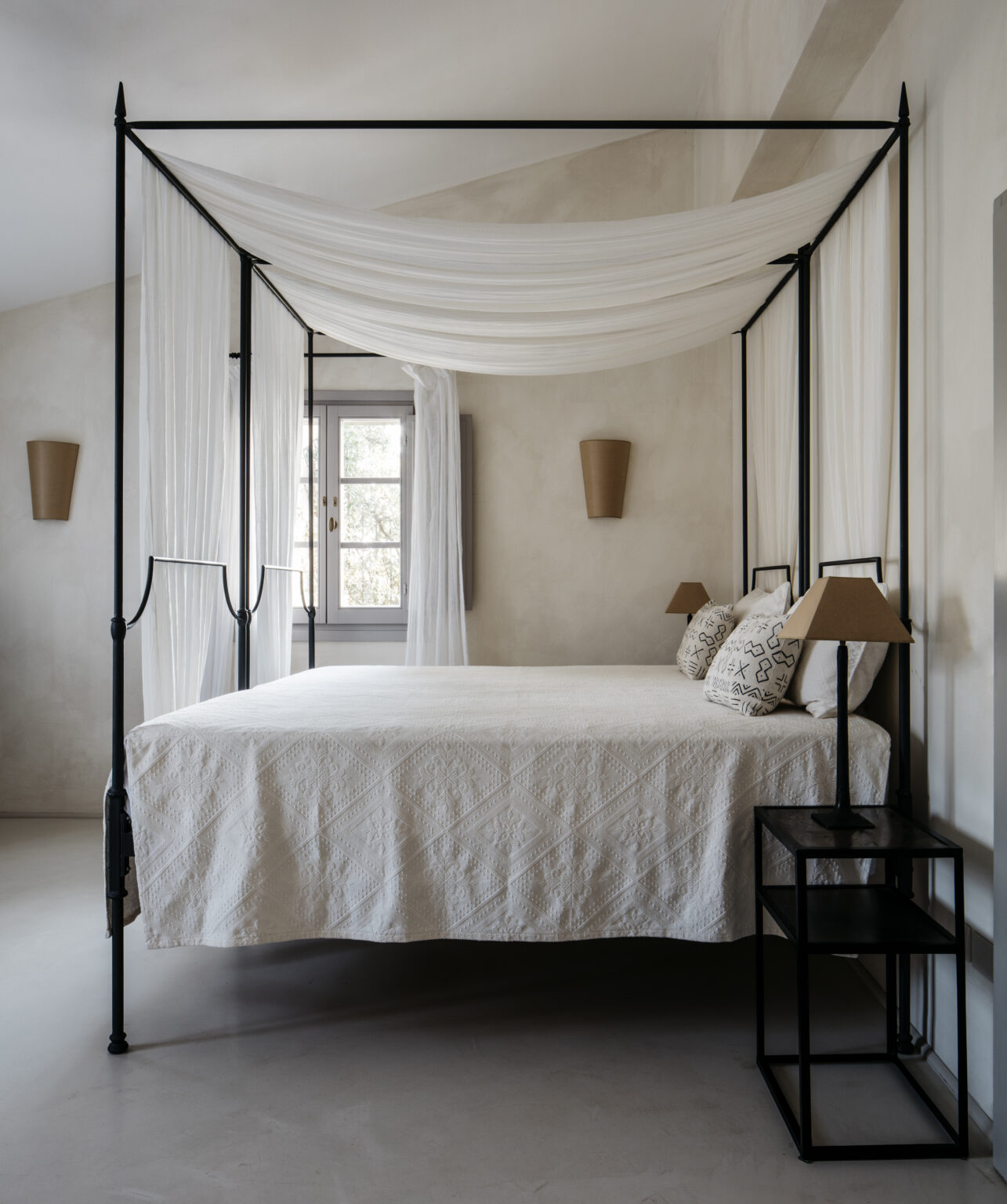 Elegant black metal four-poster bed with white drapery and decorative pillows in a minimalist bedroom featuring soft neutral tones and modern lighting. | Only on SopranoVillas