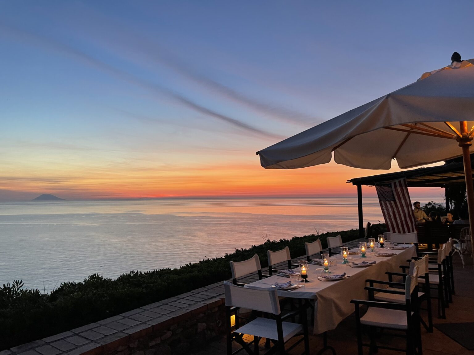 Scenic sunset view from a coastal restaurant with a candlelit dining setup, overlooking calm waters and distant mountains. The sky displays vibrant hues of orange, pink, and blue, creating a romantic atmosphere for diners. | Only on SopranoVillas