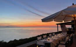 Scenic sunset view from a coastal restaurant with a candlelit dining setup, overlooking calm waters and distant mountains. The sky displays vibrant hues of orange, pink, and blue, creating a romantic atmosphere for diners.