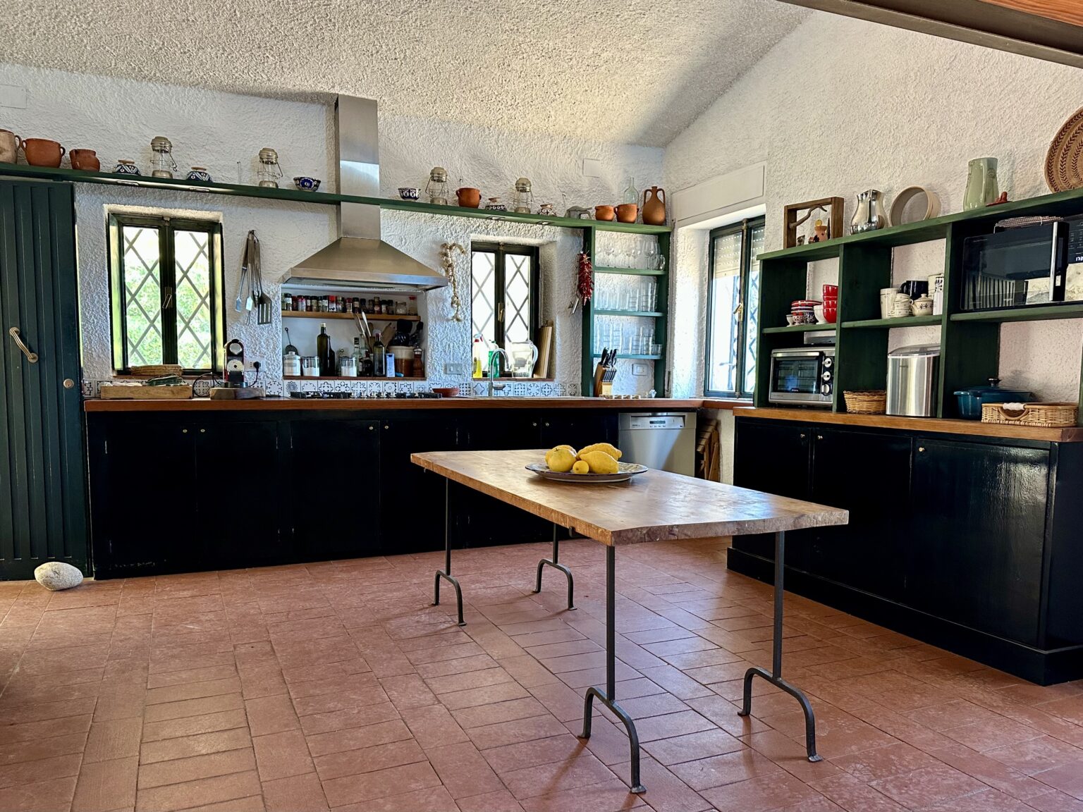 Bright and spacious kitchen featuring a central wooden table with lemons, black cabinets, and green shelves filled with dishes and cooking essentials. The room is illuminated by natural light from the windows, showcasing a blend of rustic and modern design elements. | Only on SopranoVillas