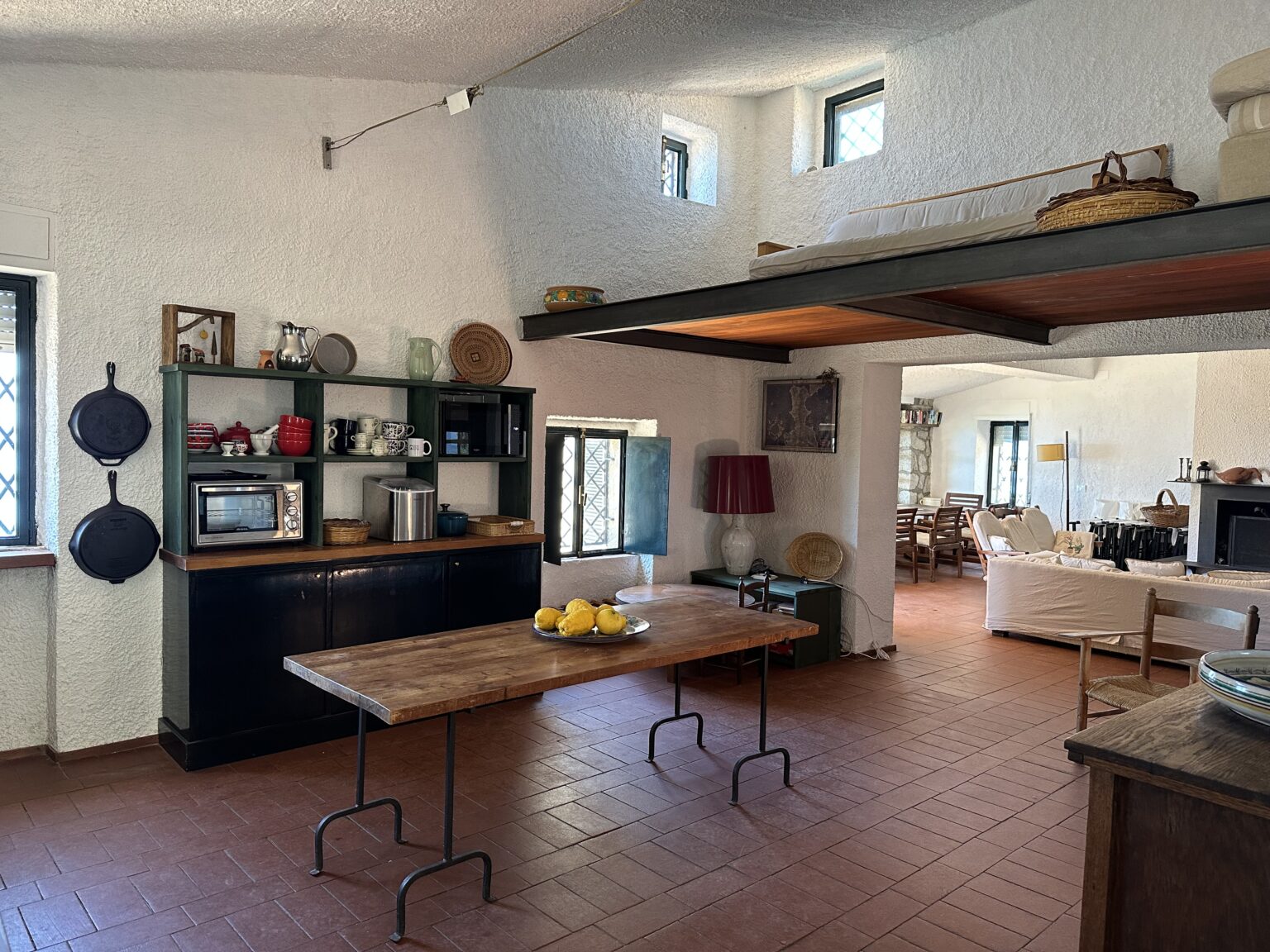 Bright and airy kitchen with a rustic design featuring a wooden table, open shelving with kitchenware, and a cozy living area visible in the background. The space has terracotta flooring, white textured walls, and large windows allowing natural light to flood in. | Only on SopranoVillas