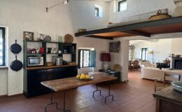 Bright and airy kitchen with a rustic design featuring a wooden table, open shelving with kitchenware, and a cozy living area visible in the background. The space has terracotta flooring, white textured walls, and large windows allowing natural light to flood in.