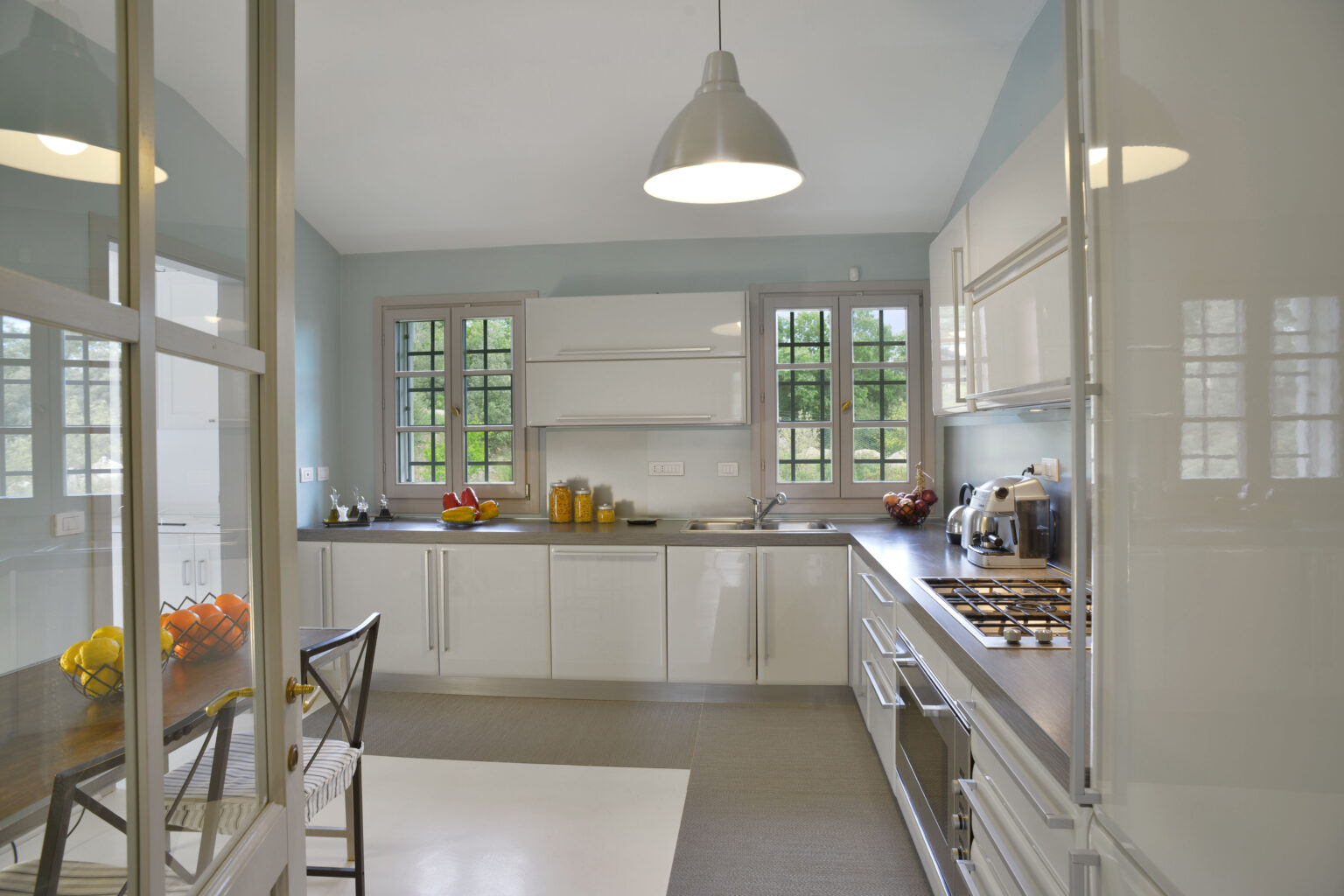 Modern kitchen interior featuring sleek white cabinets, a wooden countertop, and large windows allowing natural light. The space includes a dining table with a fruit bowl, a coffee maker, and a stove, creating a bright and inviting atmosphere. | Only on SopranoVillas