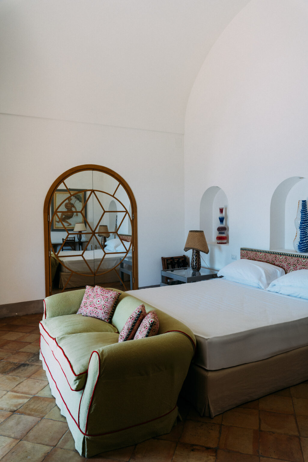 Modern bedroom interior featuring a large bed with decorative pillows, a stylish lamp, and a geometric mirror reflecting the room's design elements. The space is characterized by white walls and elegant decor, creating a serene atmosphere. | Only on SopranoVillas