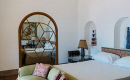 Modern bedroom interior featuring a large bed with decorative pillows, a stylish lamp, and a geometric mirror reflecting the room's design elements. The space is characterized by white walls and elegant decor, creating a serene atmosphere.