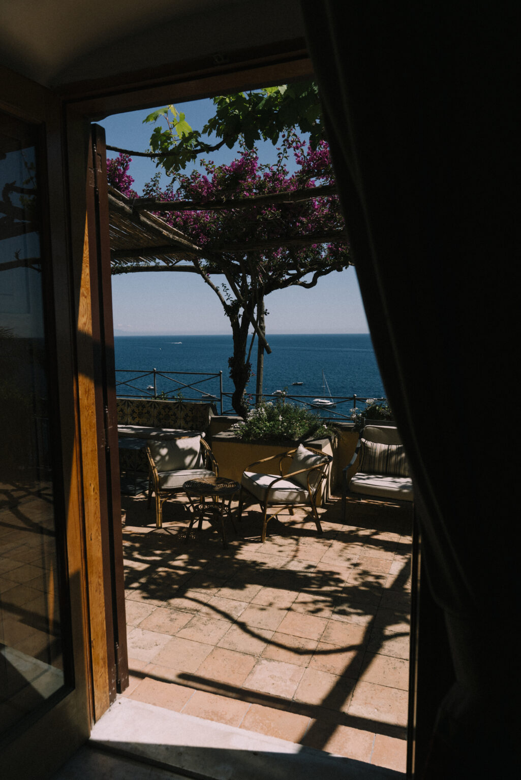 View from a doorway showcasing a cozy outdoor seating area with plants, overlooking a serene blue ocean and clear sky. | Only on SopranoVillas
