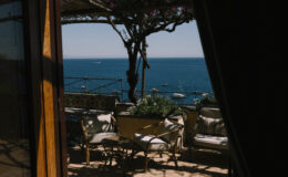 View from a doorway showcasing a cozy outdoor seating area with plants, overlooking a serene blue ocean and clear sky.
