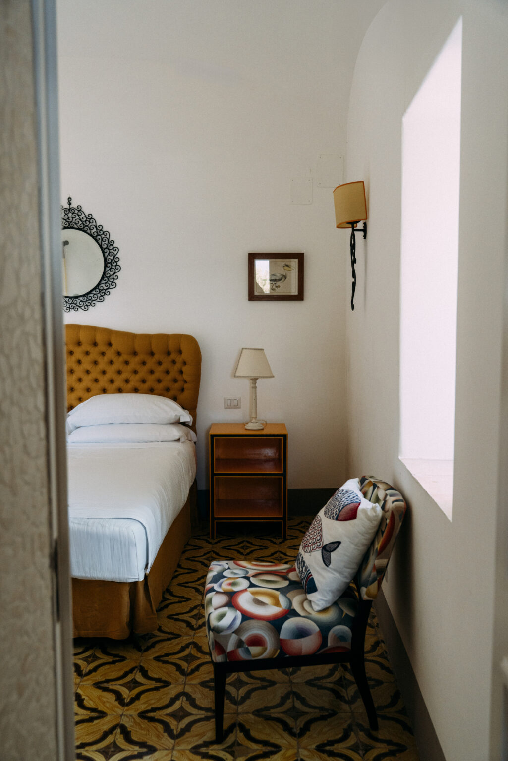 Cozy bedroom featuring a yellow tufted headboard, a bedside lamp, and wooden nightstand, accented by a decorative mirror and framed artwork on the wall. Natural light illuminates the space, creating a warm and inviting atmosphere. | Only on SopranoVillas