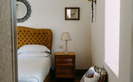 Cozy bedroom featuring a yellow tufted headboard, a bedside lamp, and wooden nightstand, accented by a decorative mirror and framed artwork on the wall. Natural light illuminates the space, creating a warm and inviting atmosphere.