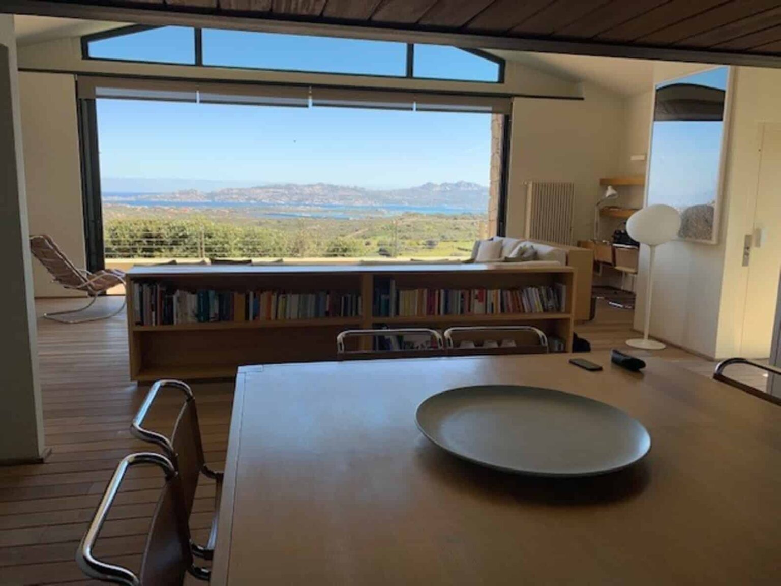 Modern interior of a bright living space featuring a wooden dining table, a large bookshelf, and panoramic windows showcasing a scenic mountain view. The design emphasizes natural light and open space, creating a serene atmosphere. | Only on SopranoVillas