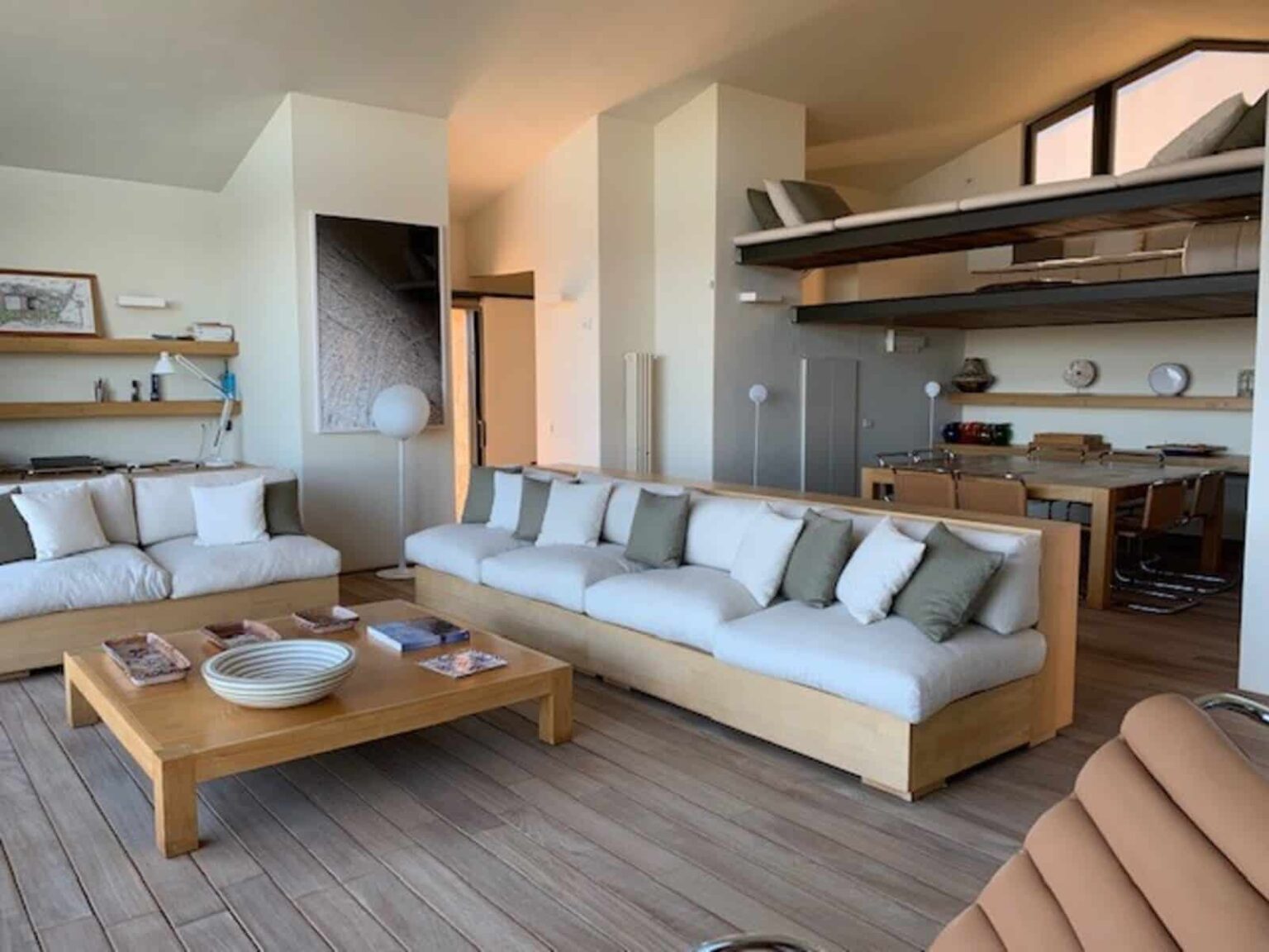 Modern living room featuring a spacious layout with a large sectional sofa adorned with neutral-colored cushions, a wooden coffee table, and minimalist decor. The room has large windows allowing natural light, a cozy dining area in the background, and wooden shelving for added storage. Ideal for contemporary home design inspiration. | Only on SopranoVillas