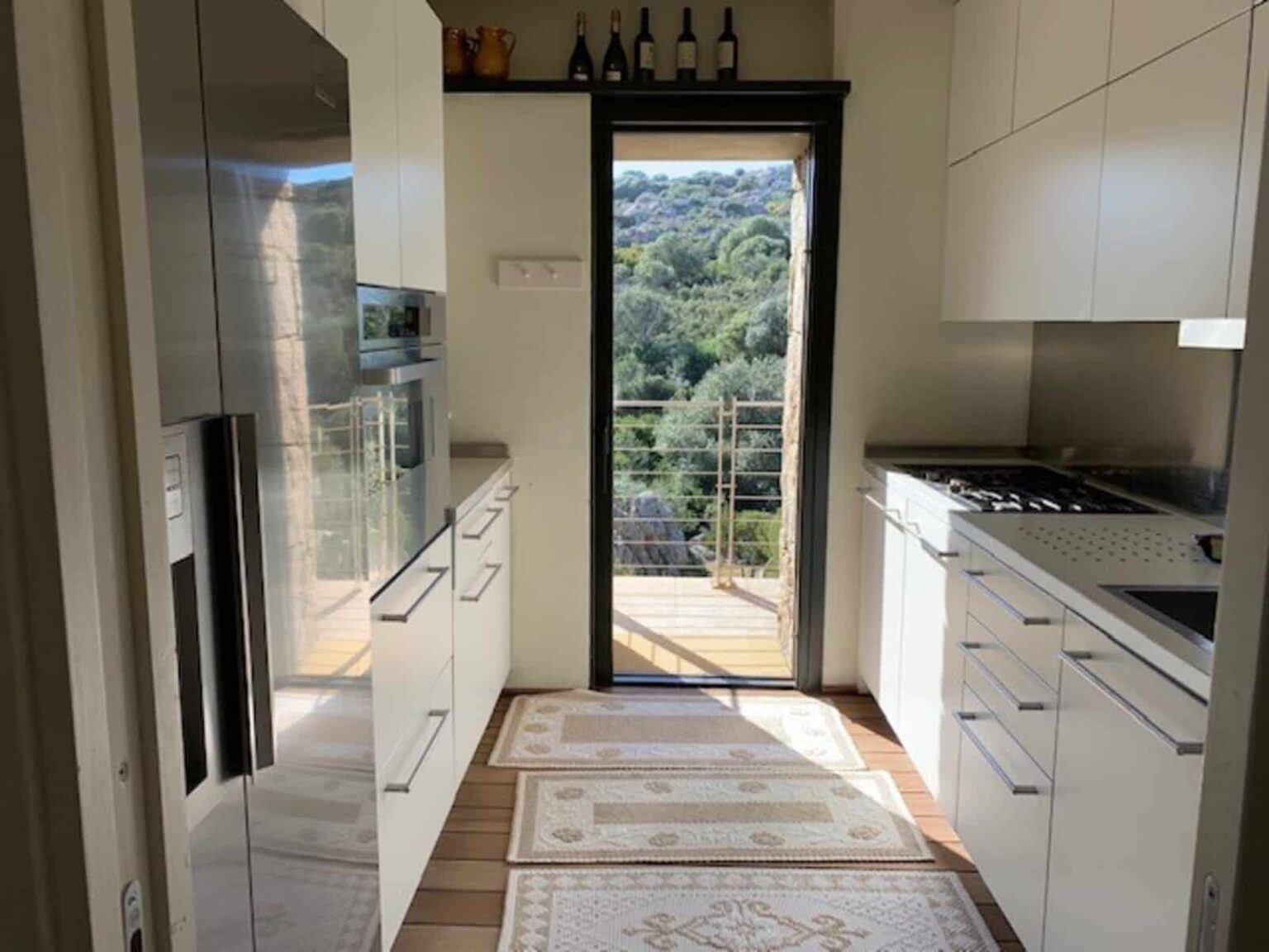 Modern kitchen interior with sleek white cabinetry, stainless steel appliances, and a view of greenery through a sliding glass door. Natural light enhances the spacious layout, featuring a stylish rug on the wooden floor. | Only on SopranoVillas