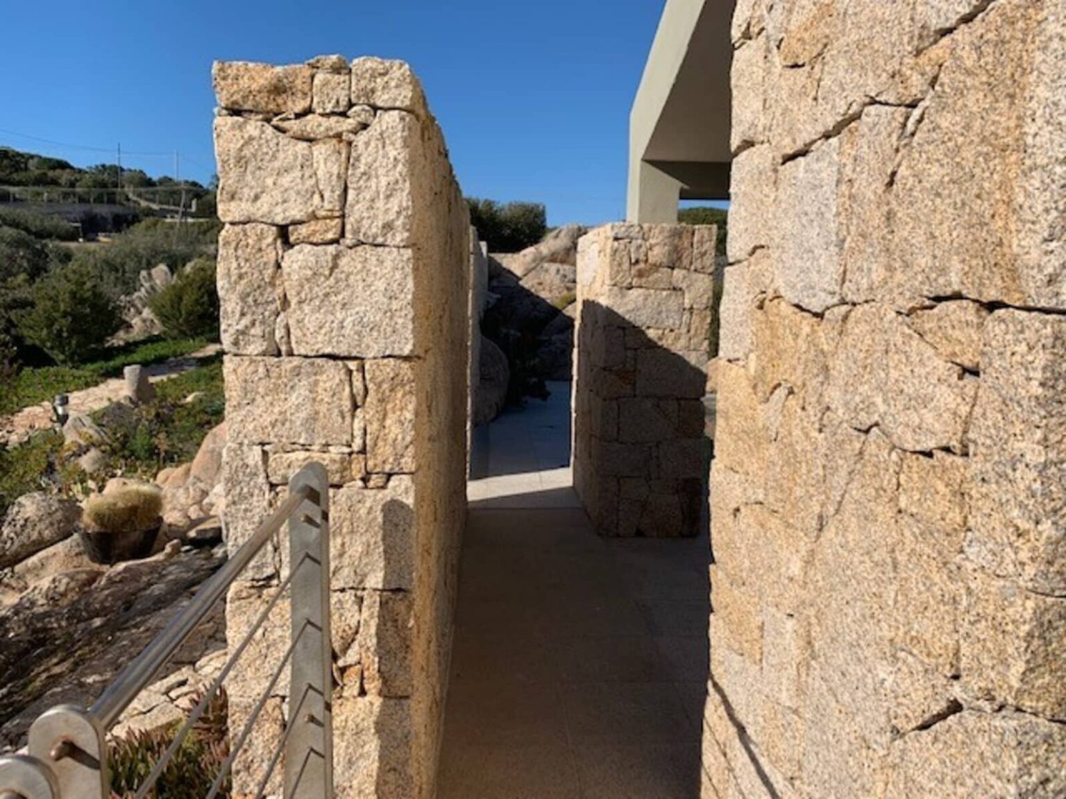 Narrow stone pathway between high walls, leading to a modern architectural structure, surrounded by greenery and clear blue skies. | Only on SopranoVillas