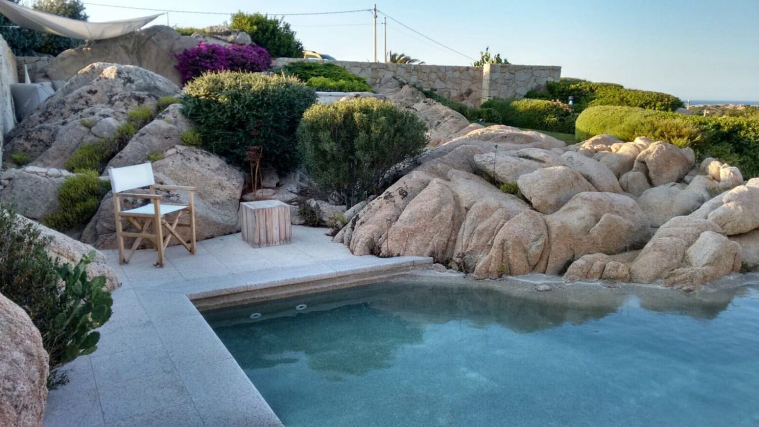 Serene outdoor pool surrounded by natural rock formations and lush greenery, featuring a wooden chair and a small table, ideal for relaxation in a tranquil setting. | Only on SopranoVillas