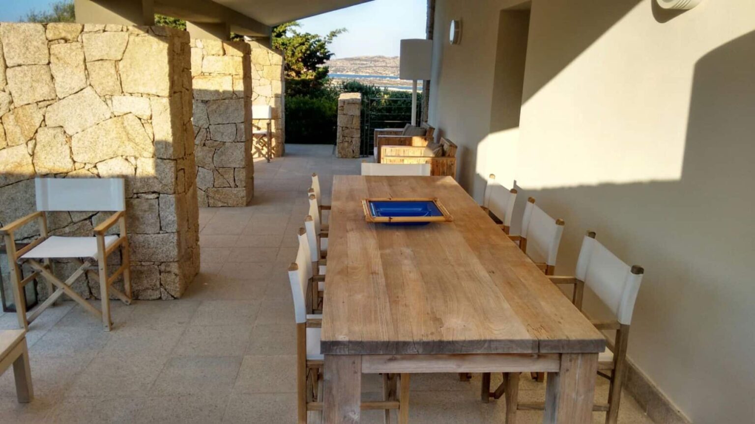 Outdoor dining area featuring a large wooden table and white chairs, surrounded by a stone wall and a scenic view. Ideal for al fresco meals and gatherings. | Only on SopranoVillas